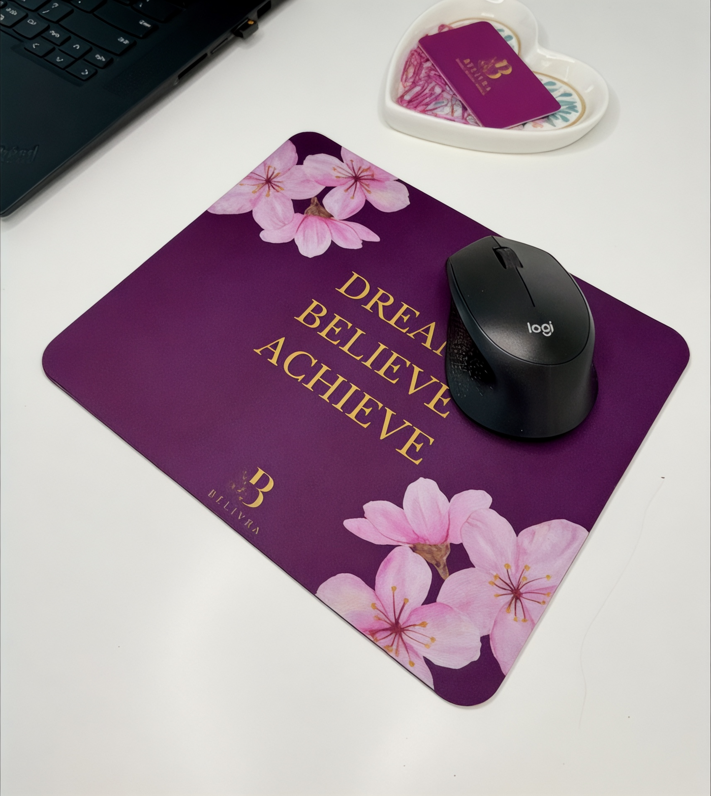 Belivra cherry blossom mouse pad with gold motivational quote "Dream Believe Achieve" – shown on a white desk with a black Logitech mouse, laptop, and heart-shaped tray. Feminine and empowering desk decor for women entrepreneurs, students, or home office inspiration.