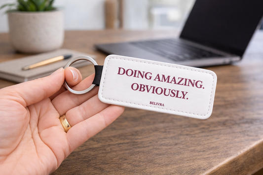 Hand holding a Belivra keychain with the text “Doing amazing. Obviously.” on a desk with a laptop and plant in the background.