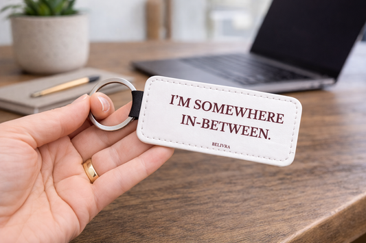 Hand holding a Belivra keychain with the text “I'm somewhere in-between.” on a desk with a laptop and plant in the background.
