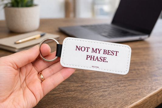Hand holding a Belivra keychain with the text “Not my best phase.” on a desk with a laptop and plant in the background.