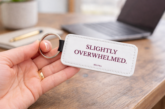 Hand holding a Belivra keychain with the text “Slightly overwhelmed.” on a desk with a laptop and plant in the background.