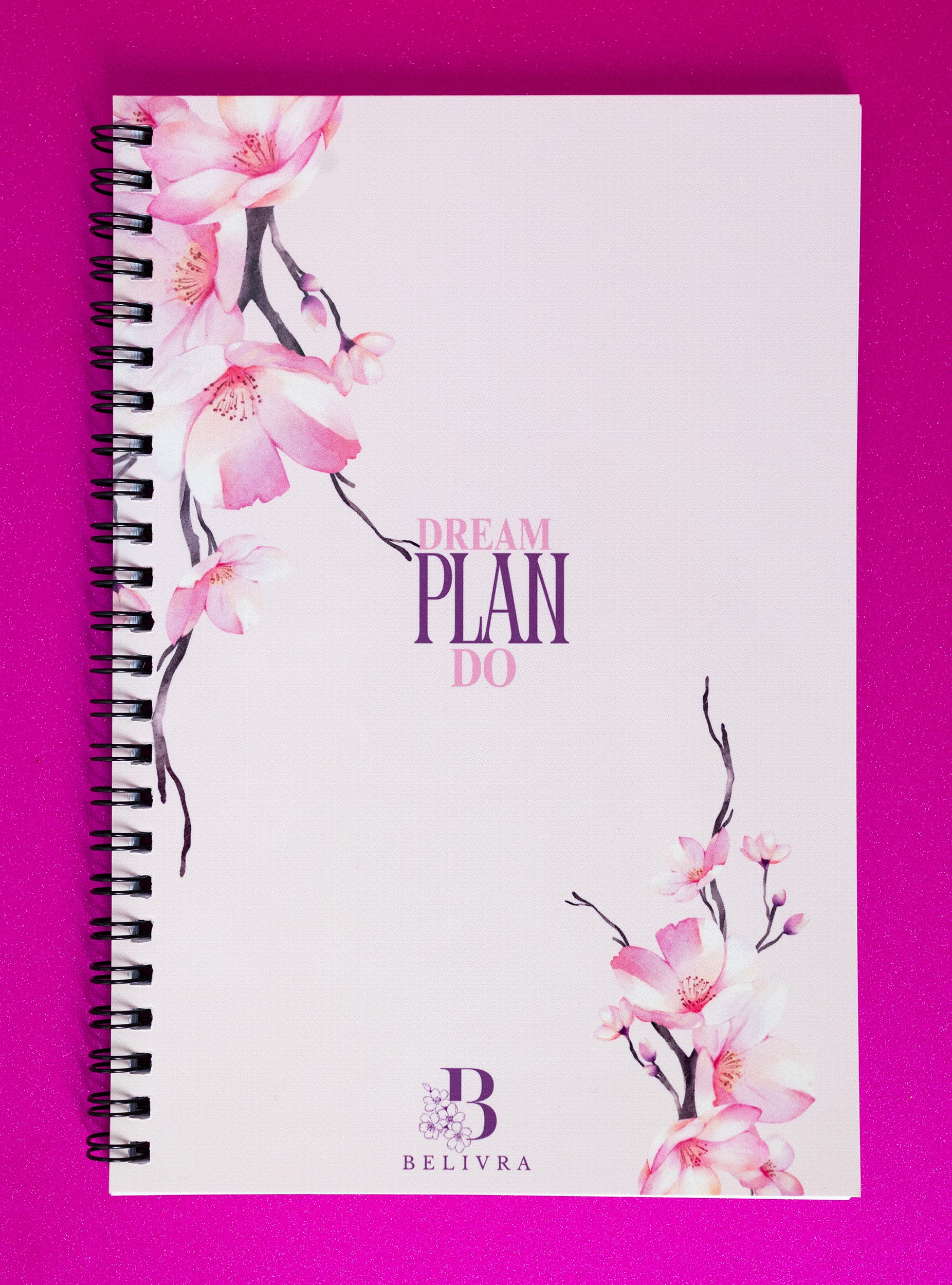 Spiral-bound notebook with a soft white cover adorned with pink magnolia flowers along the left and right edges. The motivational phrase “DREAM PLAN DO” is displayed in elegant purple font, positioned at the center of the cover. The notebook is branded with a small “B” logo at the bottom, symbolizing Belivra. A stylish, practical tool designed to inspire action and goal setting.