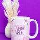 Belivra ceramic mug featuring the motivational phrase "Sip the Dream, Live the Vision" in elegant pink and purple text, surrounded by soft cherry blossom illustrations. Displayed on a vibrant purple background with a soft floral arrangement, this inspiring mug is perfect for sipping coffee, tea, or your favorite beverage while focusing on goals and dreams.
