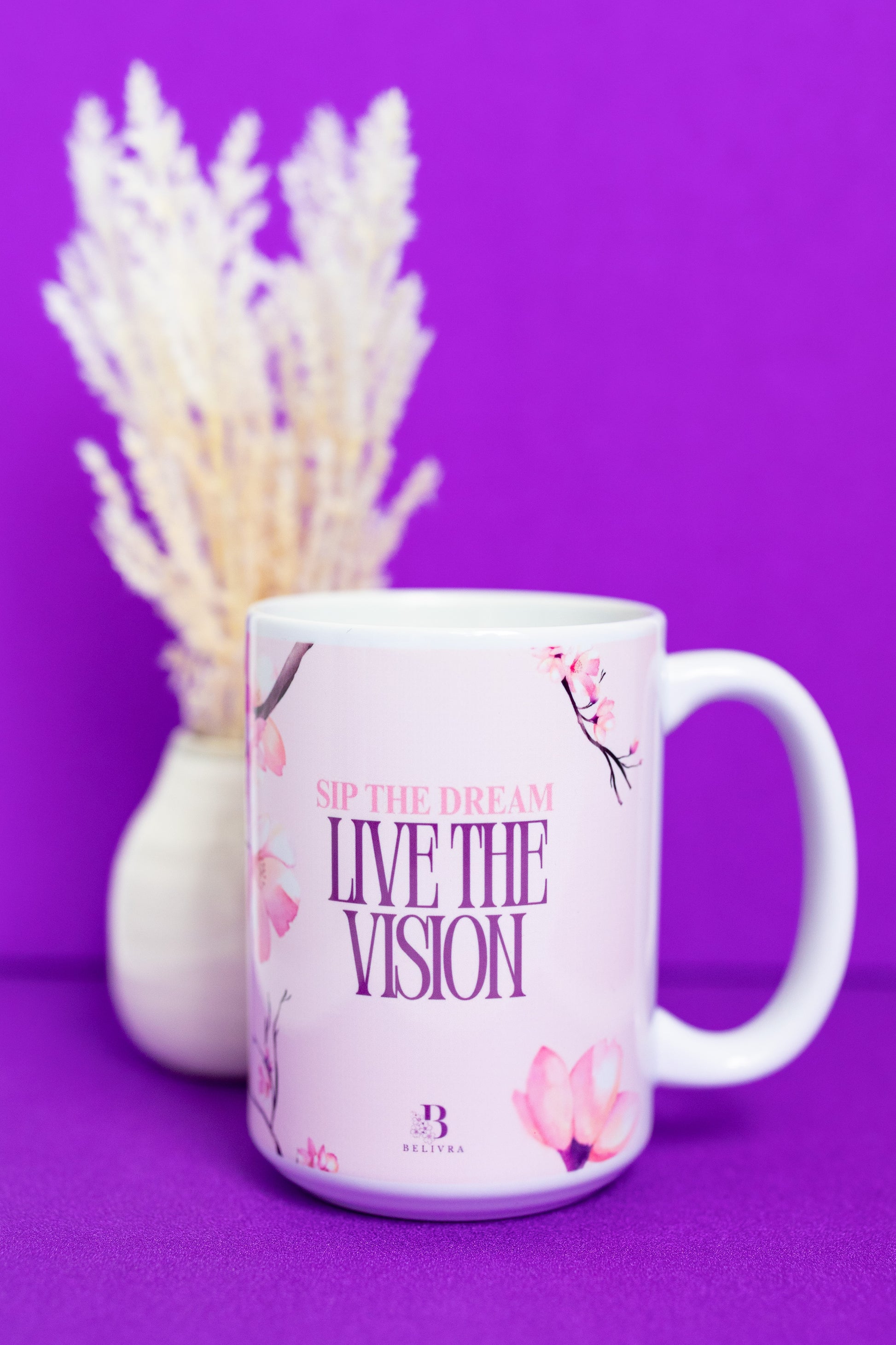 Belivra ceramic mug featuring the motivational phrase "Sip the Dream, Live the Vision" in elegant pink and purple text, surrounded by soft cherry blossom illustrations. Displayed on a vibrant purple background with a soft floral arrangement, this inspiring mug is perfect for sipping coffee, tea, or your favorite beverage while focusing on goals and dreams.