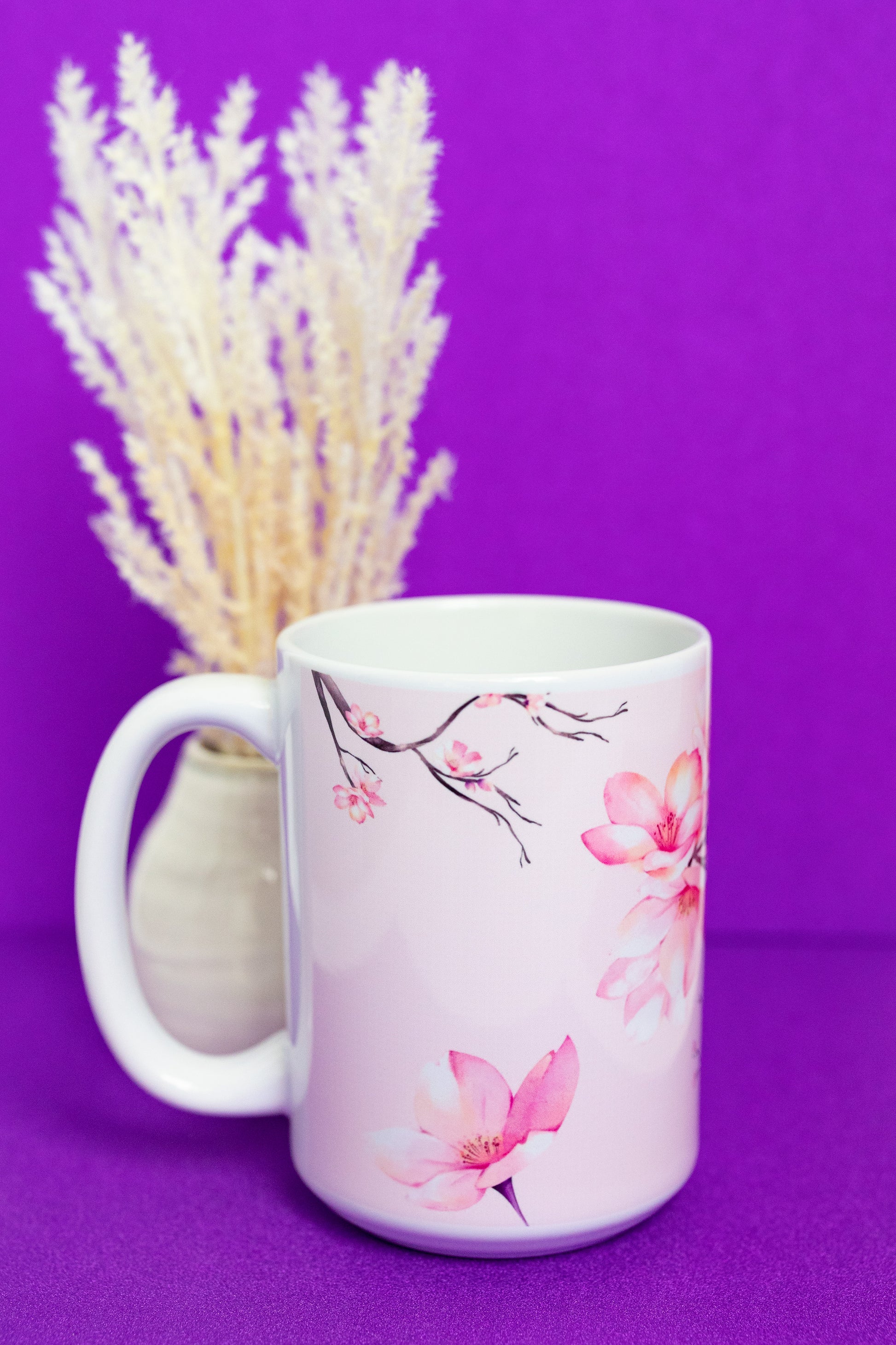 Side view of Belivra ceramic mug with soft pink cherry blossom illustrations along the handle and rim, beautifully accentuating the floral design. Set against a vibrant purple background with delicate floral stems, this elegant mug is perfect for sipping your favorite drink while embracing calm and mindfulness.