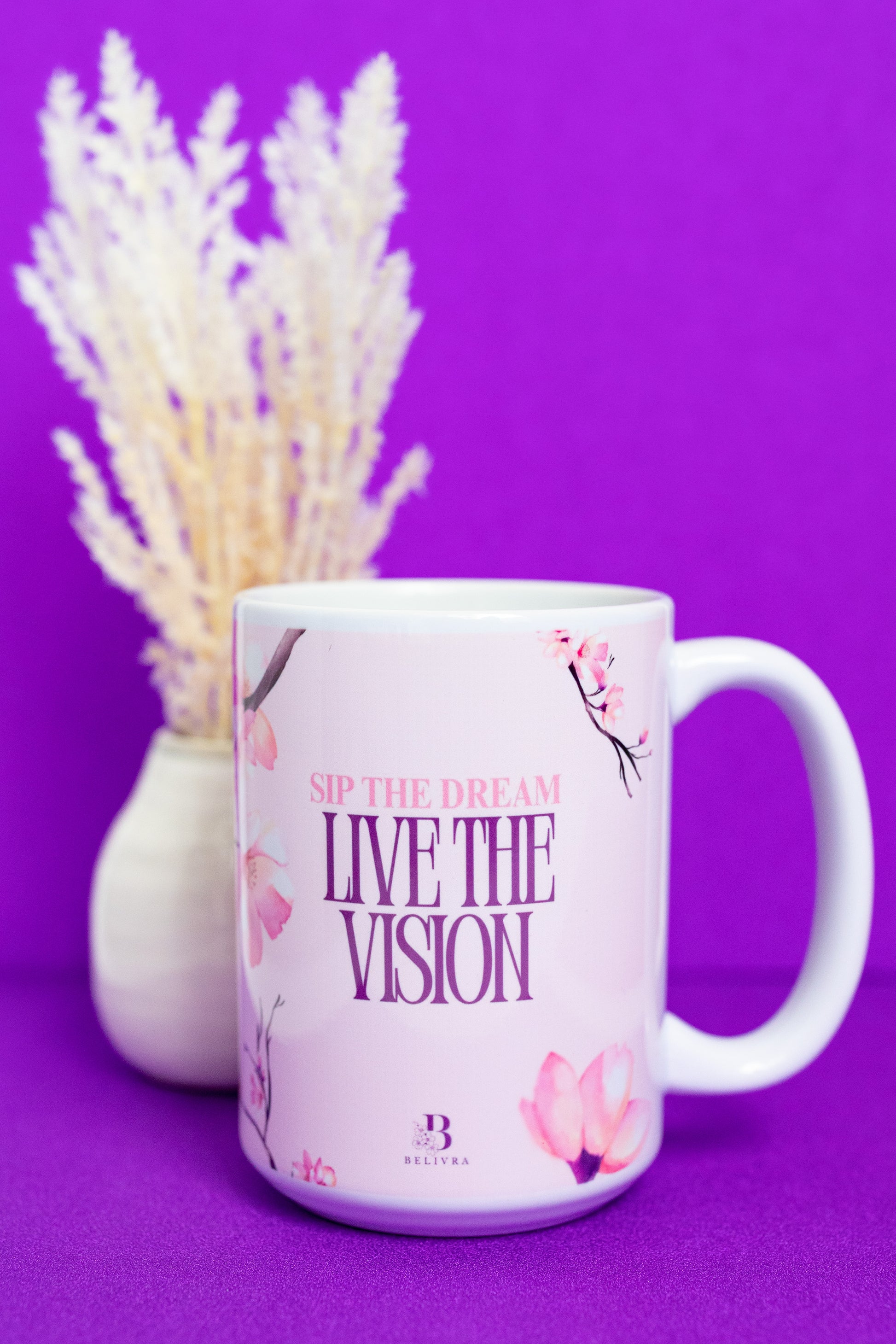 Front view of Belivra ceramic mug with the empowering phrase "Sip the Dream, Live the Vision" in elegant pink typography, surrounded by soft pink cherry blossoms. Displayed on a vibrant purple background with a delicate floral arrangement, perfect for mindful sips, positive affirmations, and adding elegance to your morning coffee routine.