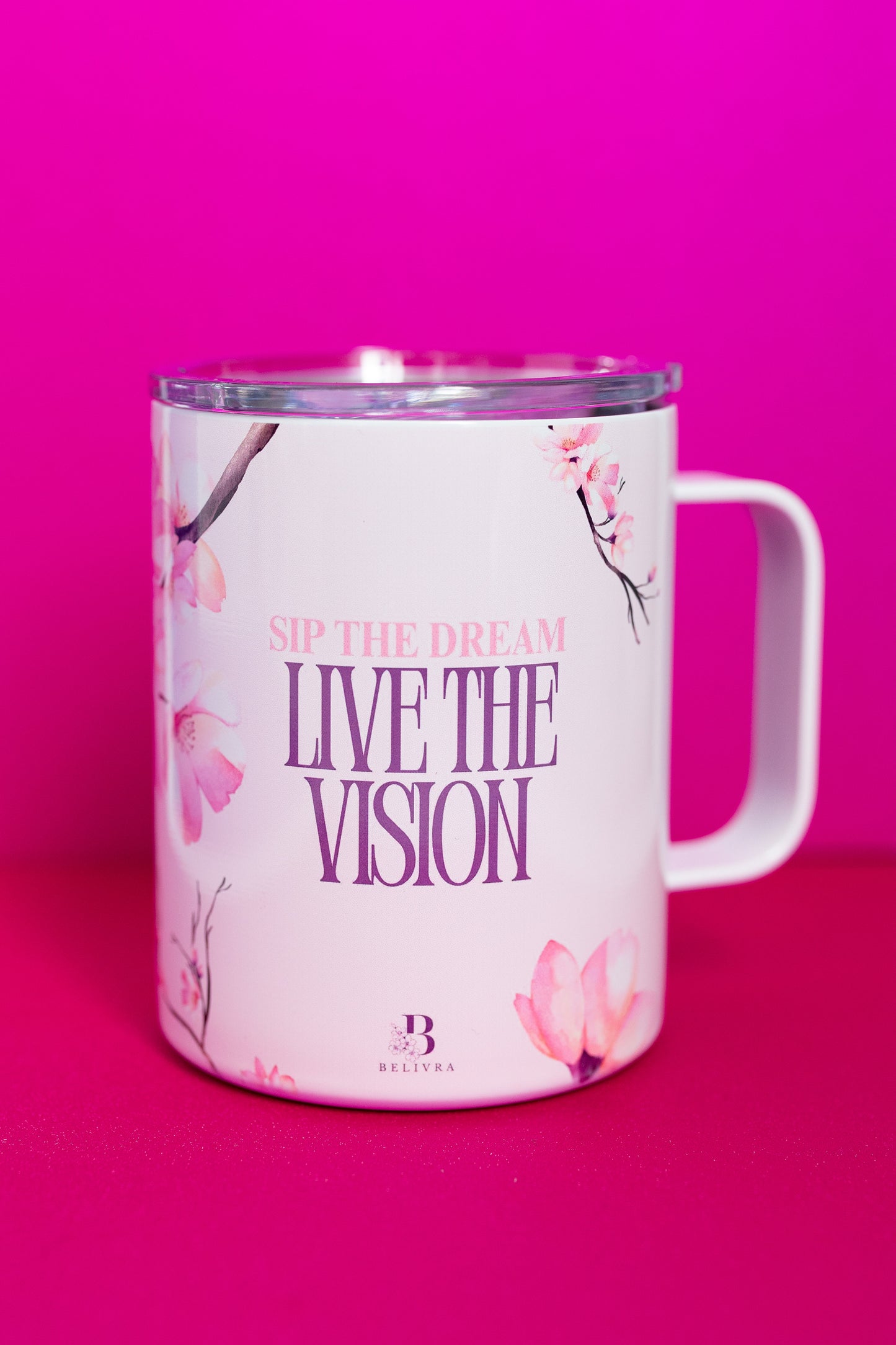 Angled view of Belivra ceramic mug with the inspirational phrase "Sip the Dream, Live the Vision" in bold purple text. The design is complemented by soft cherry blossom flowers on a pastel pink background, and the mug comes with a spill-resistant lid, ideal for enjoying your morning coffee or tea while embracing positive affirmations and motivation.