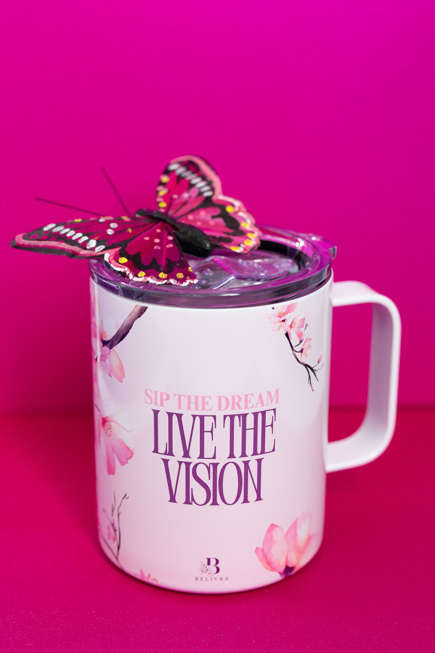A Belivra ceramic mug featuring the empowering phrase "Sip the Dream, Live the Vision" in purple text, accompanied by delicate cherry blossom designs. The mug is topped with a vibrant butterfly decoration, adding a whimsical touch. Perfect for sipping your favorite beverage while embracing positive affirmations. The mug also features a spill-resistant lid, ideal for those on the go.