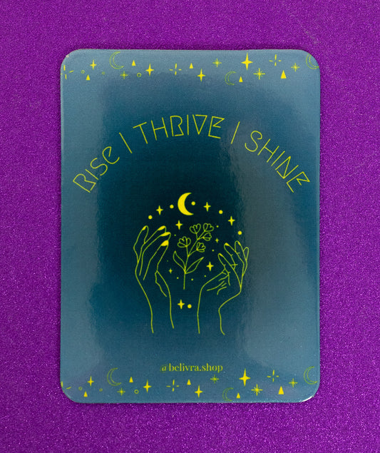 Belivra rectangular motivational magnet with the affirmation "Rise | Thrive | Shine" in bold yellow font – features celestial and floral hand illustration on a deep blue background. Displayed on a sparkly purple surface, perfect for empowering women’s workspaces, vision boards, and daily mindset reminders.