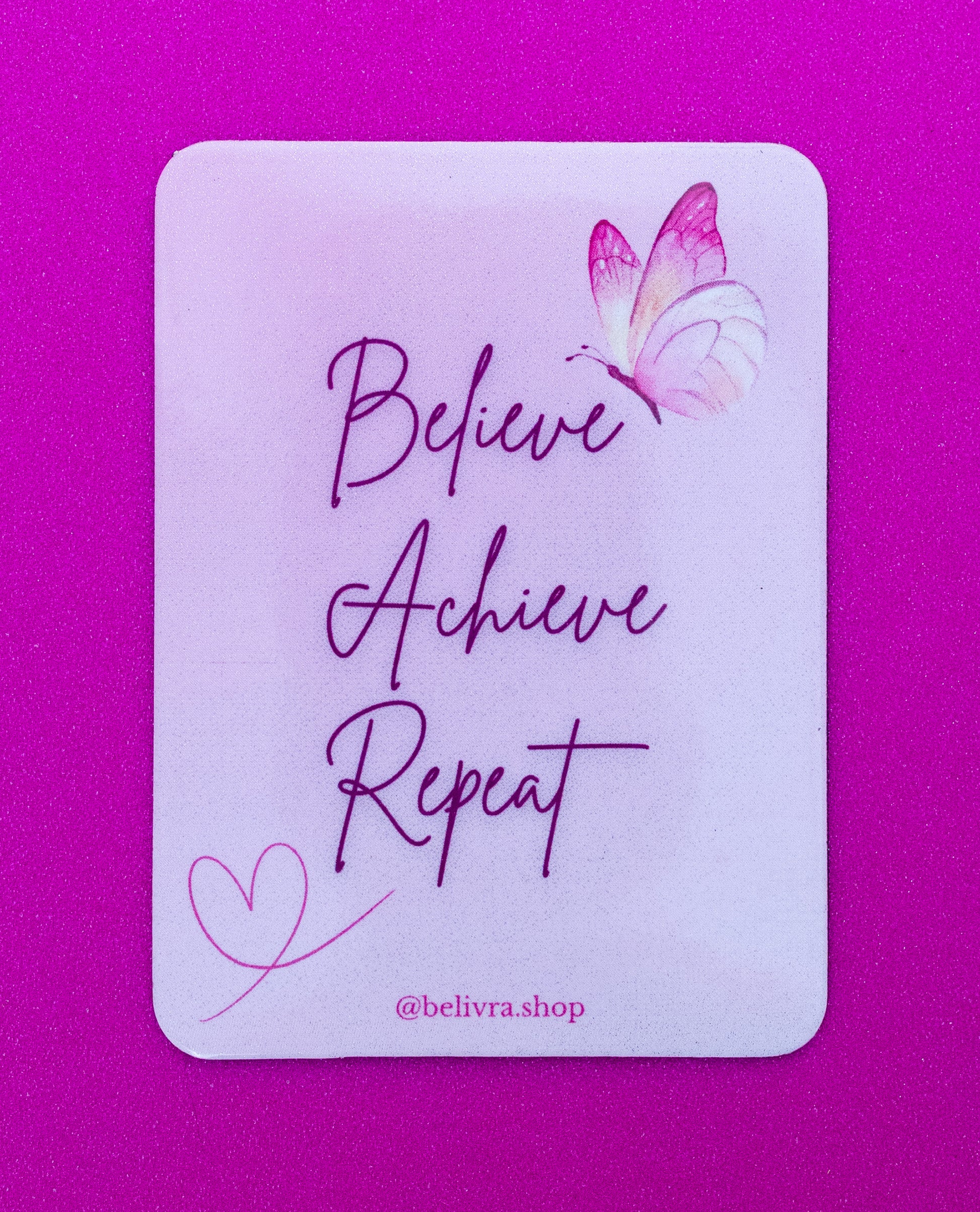Belivra rectangular motivational magnet with the quote "Believe Achieve Repeat" in elegant script font – features a pink and purple butterfly and heart doodle on a soft pastel background. Displayed on a vibrant pink surface, perfect for empowering women’s workspaces, vision boards, or self-growth reminders.