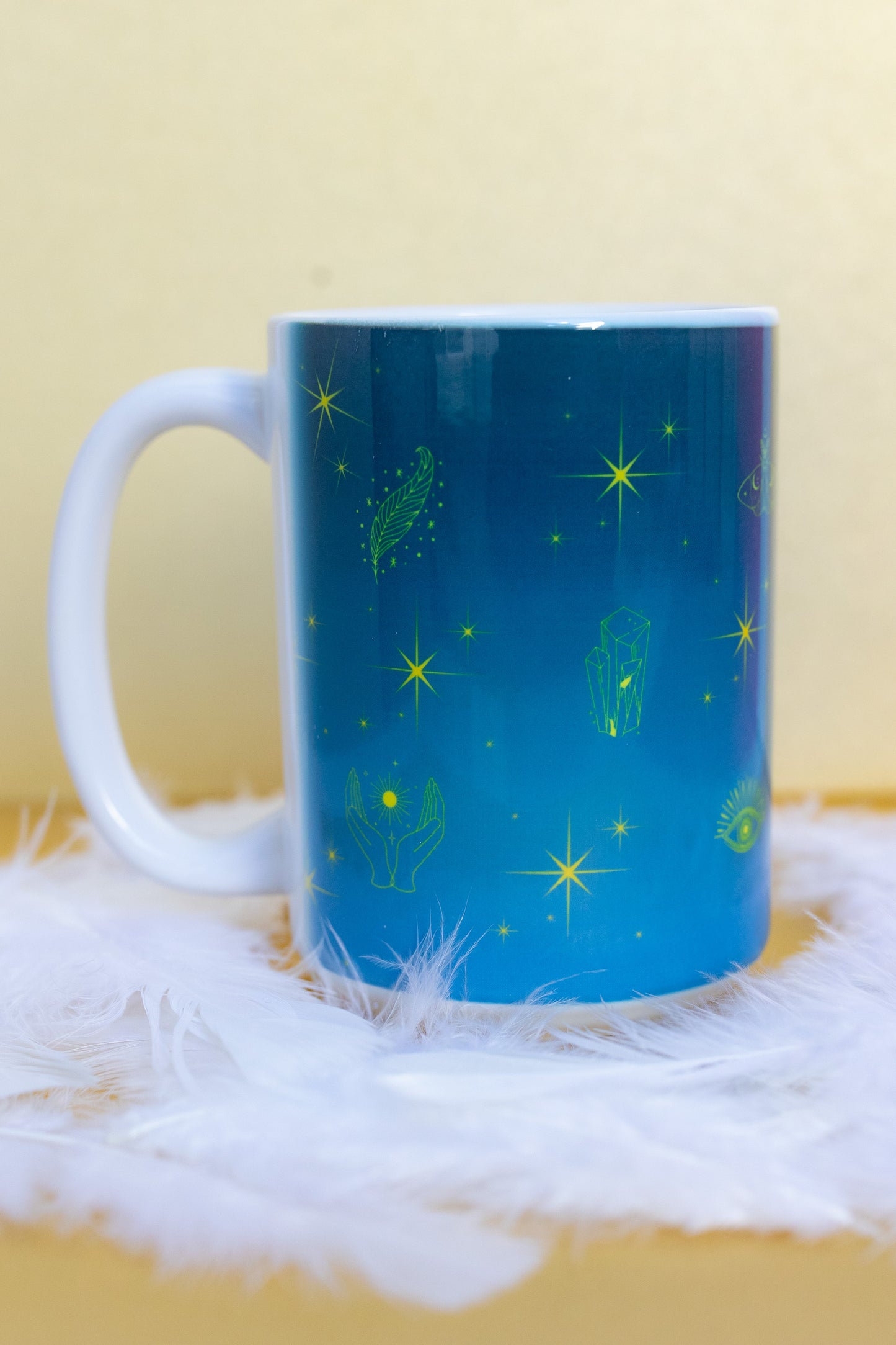 Back view of Belivra ceramic mug with a dreamy blue gradient design featuring mystical yellow illustrations – including stars, crystals, open hands, a feather, and third-eye symbols. Displayed on a soft white feather surface, perfect for spiritual morning rituals, cosmic coffee moments, and empowering gifts for mindful women and starry-eyed dreamers.