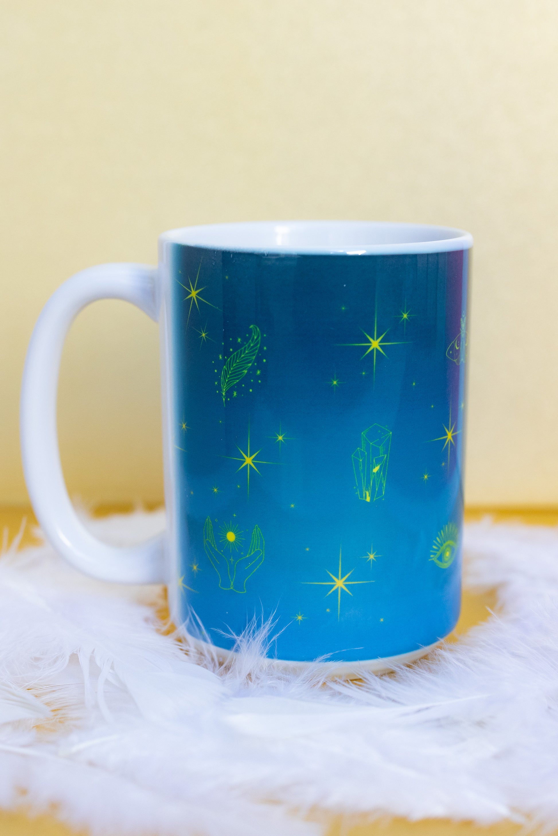 Belivra ceramic mug with a vibrant blue celestial design featuring yellow mystical illustrations like stars, open hands, feathers, crystals, and third-eye symbols. Displayed on a fluffy white feather surface, this magical coffee mug is perfect for spiritual morning rituals, mindful sipping, and cosmic desk decor for intuitive and empowered women.