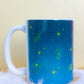 Centered back view of Belivra ceramic mug with a cosmic blue background and mystical yellow illustrations including stars, feathers, open hands, crystals, and a third-eye symbol. Displayed on a soft white feather base, this enchanting mug is ideal for spiritual morning rituals, meditation companions, and empowering gifts for astrology and energy lovers.