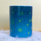 Angled back view of Belivra ceramic mug with a deep blue celestial design featuring yellow mystical illustrations like a moth, crystal, third-eye symbol, and stars. Displayed on a fluffy white feather surface, this magical and empowering mug is perfect for spiritual morning routines, cosmic energy lovers, and daily coffee rituals with intention.