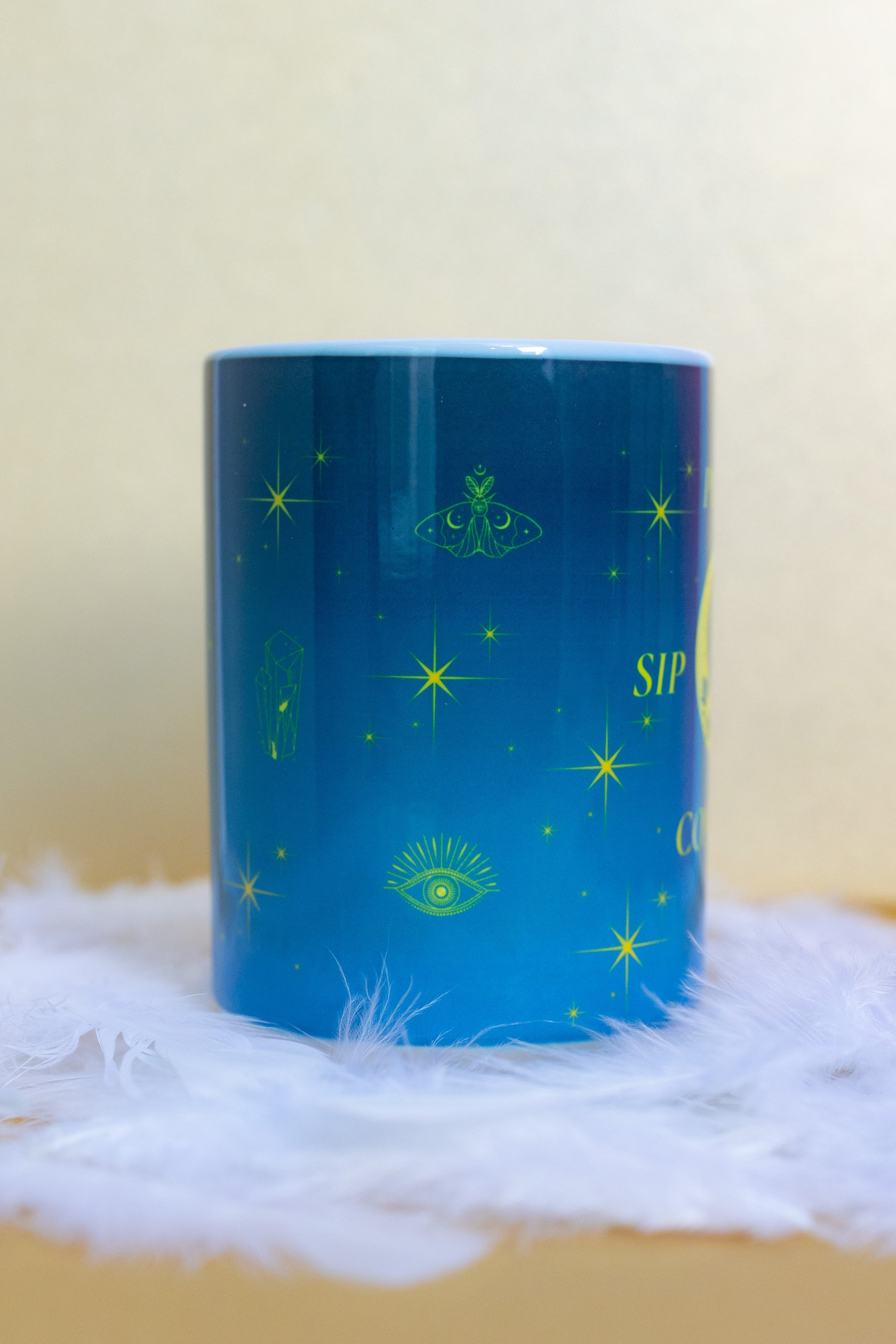 Angled back view of Belivra ceramic mug with a deep blue celestial design featuring yellow mystical illustrations like a moth, crystal, third-eye symbol, and stars. Displayed on a fluffy white feather surface, this magical and empowering mug is perfect for spiritual morning routines, cosmic energy lovers, and daily coffee rituals with intention.