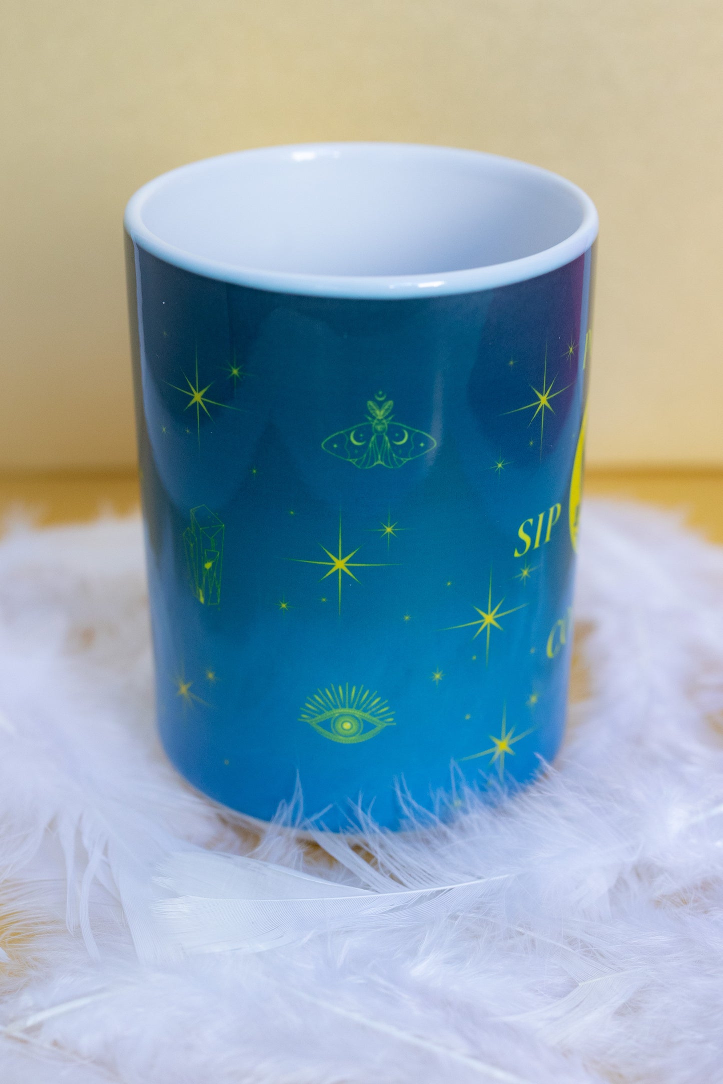 Side view of Belivra ceramic mug with a mystical blue and yellow design featuring celestial symbols like stars, crystals, a moth, third-eye, and moon elements. Styled on a soft white feather surface, this magical mug is perfect for spiritual morning routines, calming coffee moments, and empowering desk decor for dreamers and mindful women.