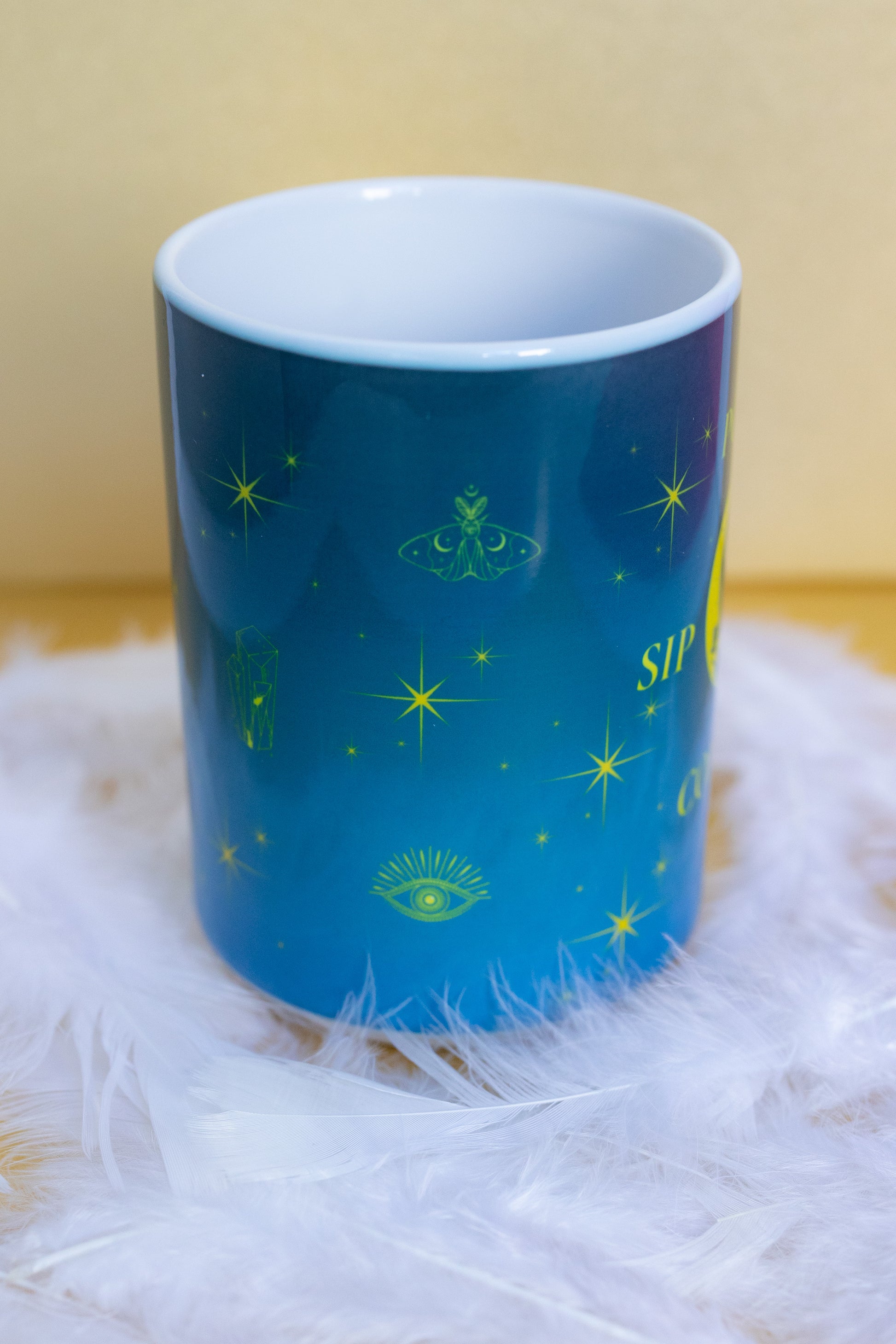 Side view of Belivra ceramic mug with a mystical blue and yellow design featuring celestial symbols like stars, crystals, a moth, third-eye, and moon elements. Styled on a soft white feather surface, this magical mug is perfect for spiritual morning routines, calming coffee moments, and empowering desk decor for dreamers and mindful women.