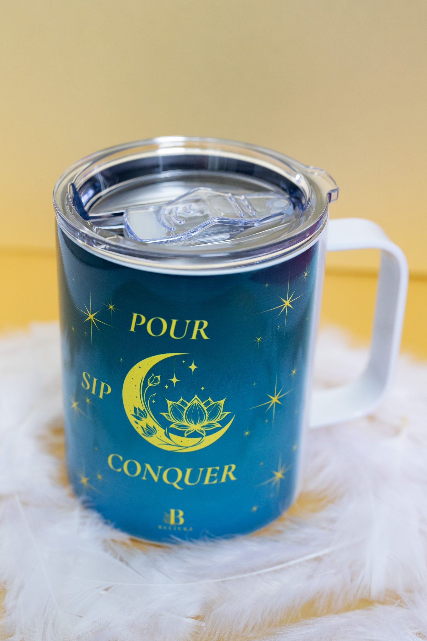 Angled view of Belivra ceramic mug with the quote "Pour. Sip. Conquer." in bold yellow text, surrounded by celestial motifs like a crescent moon, lotus, and stars, on a deep blue background. Displayed with a clear, spill-resistant lid, perfect for on-the-go coffee, tea, and empowering drinks. Ideal for morning rituals, meditation, and inspiring desk decor for spiritual and mindful women.