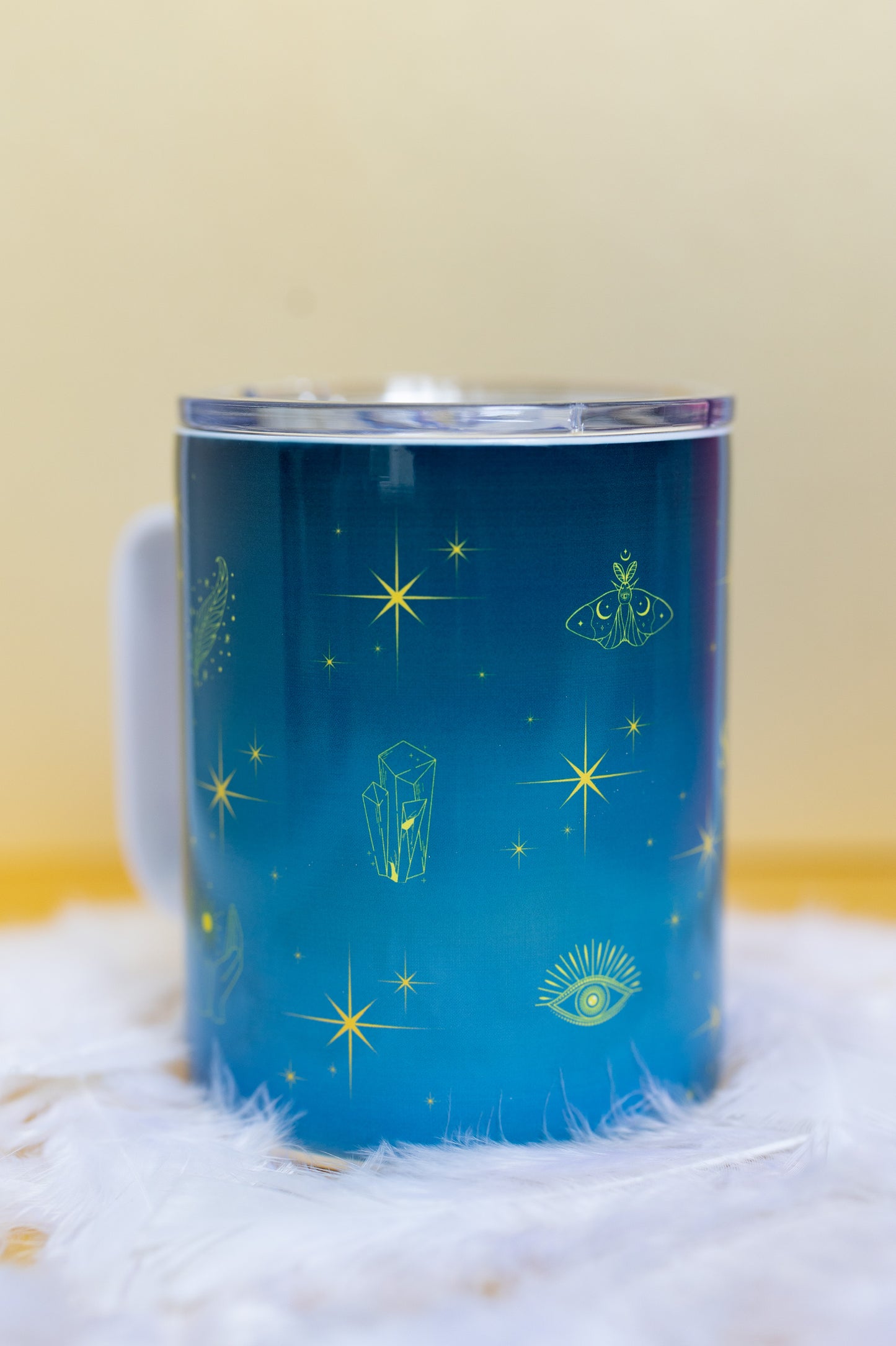 Angled side view of Belivra ceramic mug with mystical blue design featuring celestial symbols like stars, crystals, a moth, and third-eye imagery in vibrant yellow. The mug is displayed with a clear, spill-resistant lid, making it perfect for secure sipping on-the-go. Ideal for spiritual morning routines, tea lovers, and empowered women seeking mindful moments throughout their day.