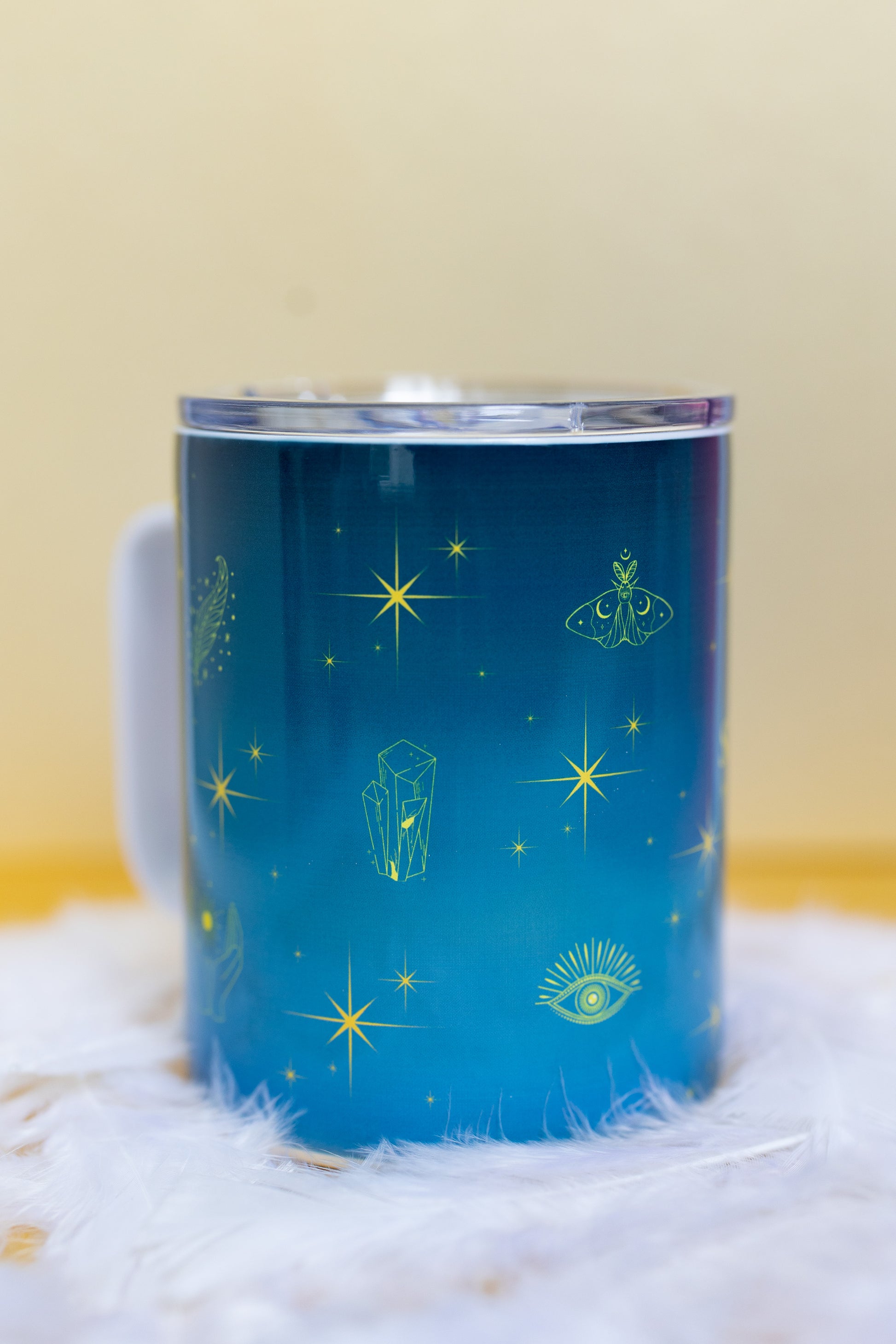 Angled side view of Belivra ceramic mug with mystical blue design featuring celestial symbols like stars, crystals, a moth, and third-eye imagery in vibrant yellow. The mug is displayed with a clear, spill-resistant lid, making it perfect for secure sipping on-the-go. Ideal for spiritual morning routines, tea lovers, and empowered women seeking mindful moments throughout their day.