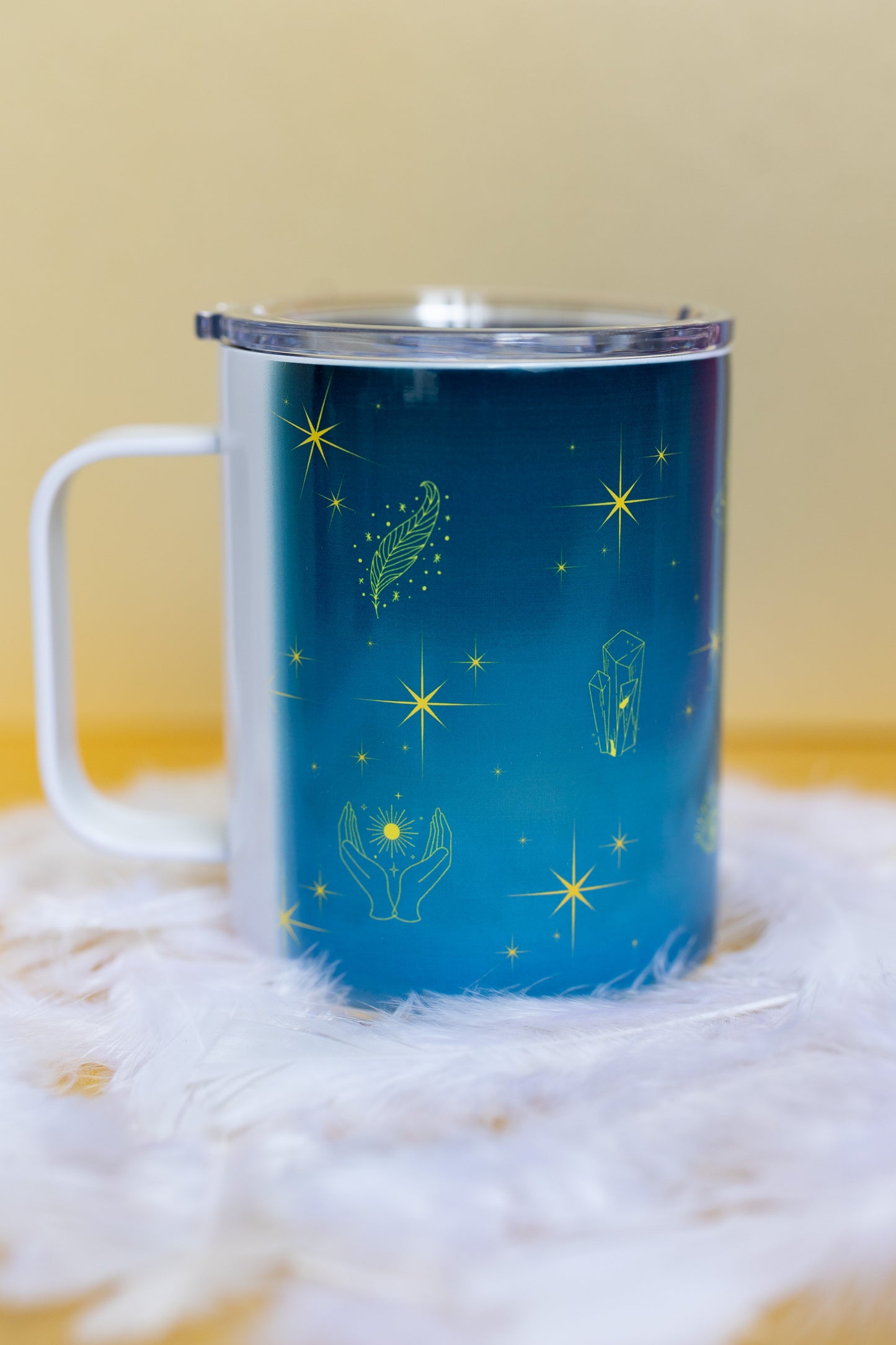 Angled view of Belivra ceramic mug with celestial blue background, featuring glowing yellow symbols like a feather, hands, crystals, and stars. The mug is shown with a spill-resistant clear lid, ideal for enjoying tea, coffee, or other beverages while embracing mindfulness. Perfect for empowered women, spiritual routines, and cozy moments.