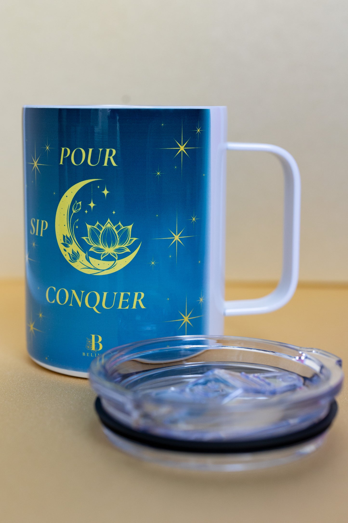 Angled view of Belivra ceramic mug with the quote "Pour. Sip. Conquer." in yellow text on a celestial blue background with a crescent moon, lotus, and stars. The mug is shown with a clear lid, ideal for maintaining the temperature of your favorite beverages. A perfect accessory for empowered morning rituals, mindful sipping, and cozy desk moments.