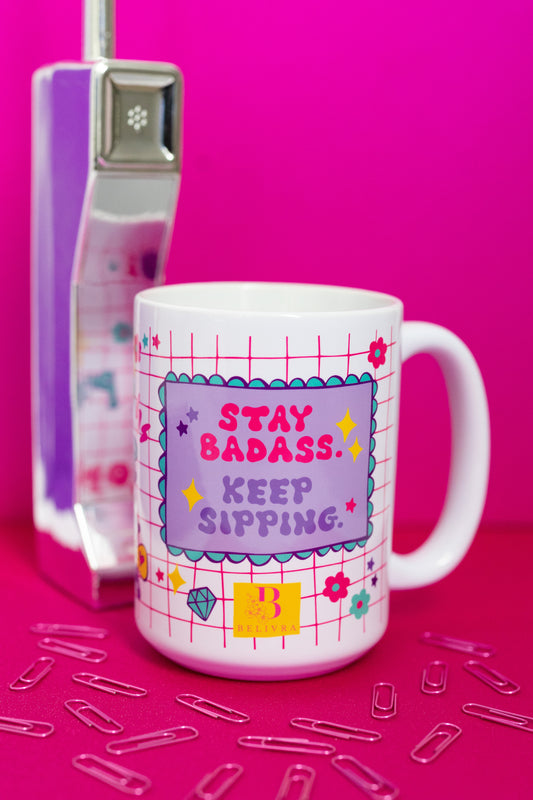 Belivra ceramic coffee mug with bold quote "Stay Badass. Keep Sipping." in playful pink and purple design – featuring a fun grid background with doodles like diamonds, gears, and stars. Displayed with a retro pink phone and scattered paperclips, this empowering mug is perfect for coffee-loving entrepreneurs, creatives, and bold women who start their day with confidence.