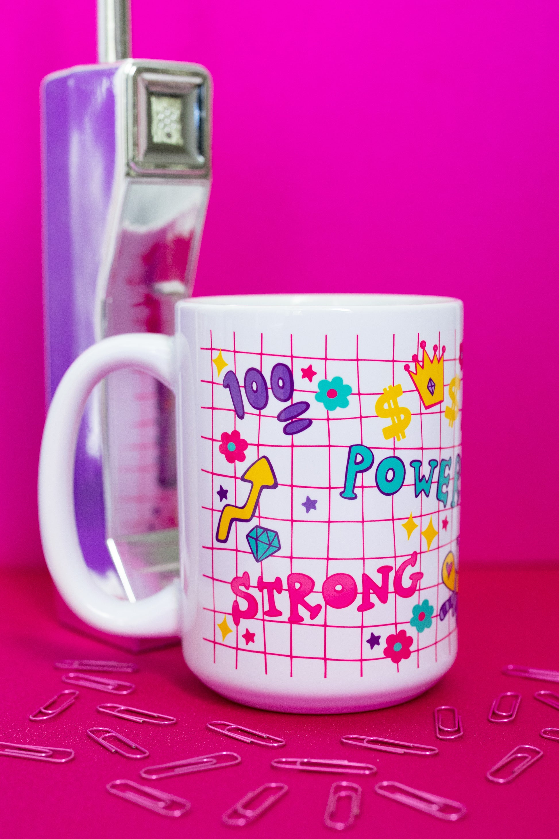 Belivra ceramic mug with colorful doodle-style design featuring empowering words like "Power" and "Strong," and fun icons like a crown, dollar signs, flexing arm, and diamonds – all on a pink grid background. Styled with a retro phone and paperclips, this bold coffee mug is perfect for confident women, office motivation, and energetic morning routines.