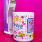 Side view of Belivra motivational coffee mug featuring colorful grid-style design with words like "Power" and "Strong," alongside playful icons like flowers, hearts, dollar signs, and a butterfly – partially showing the quote "Stay Badass. Keep Sipping." Styled with a retro phone and paperclips on a hot pink background, perfect for bold women and vibrant workspaces.
