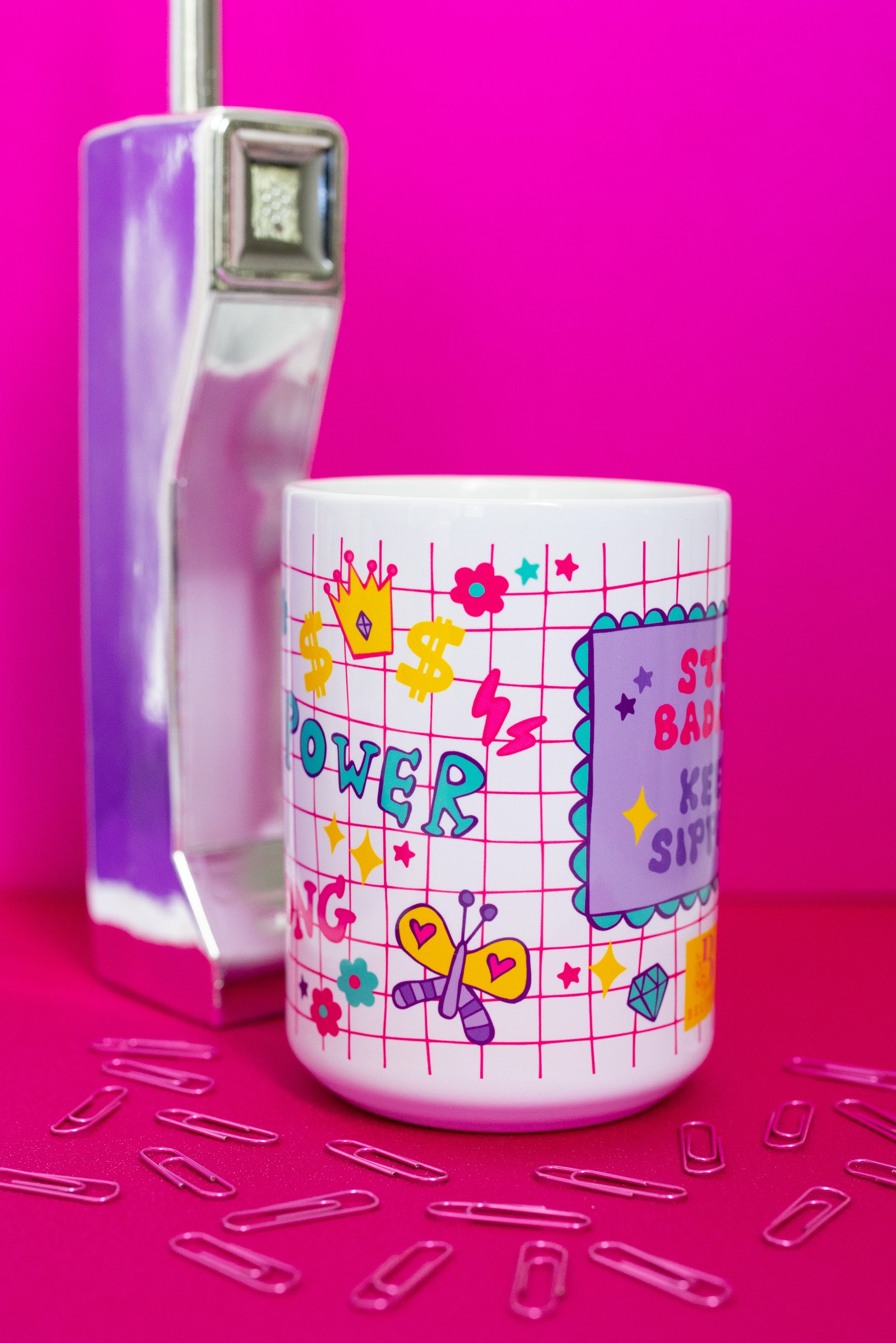Side view of Belivra motivational coffee mug featuring colorful grid-style design with words like "Power" and "Strong," alongside playful icons like flowers, hearts, dollar signs, and a butterfly – partially showing the quote "Stay Badass. Keep Sipping." Styled with a retro phone and paperclips on a hot pink background, perfect for bold women and vibrant workspaces.