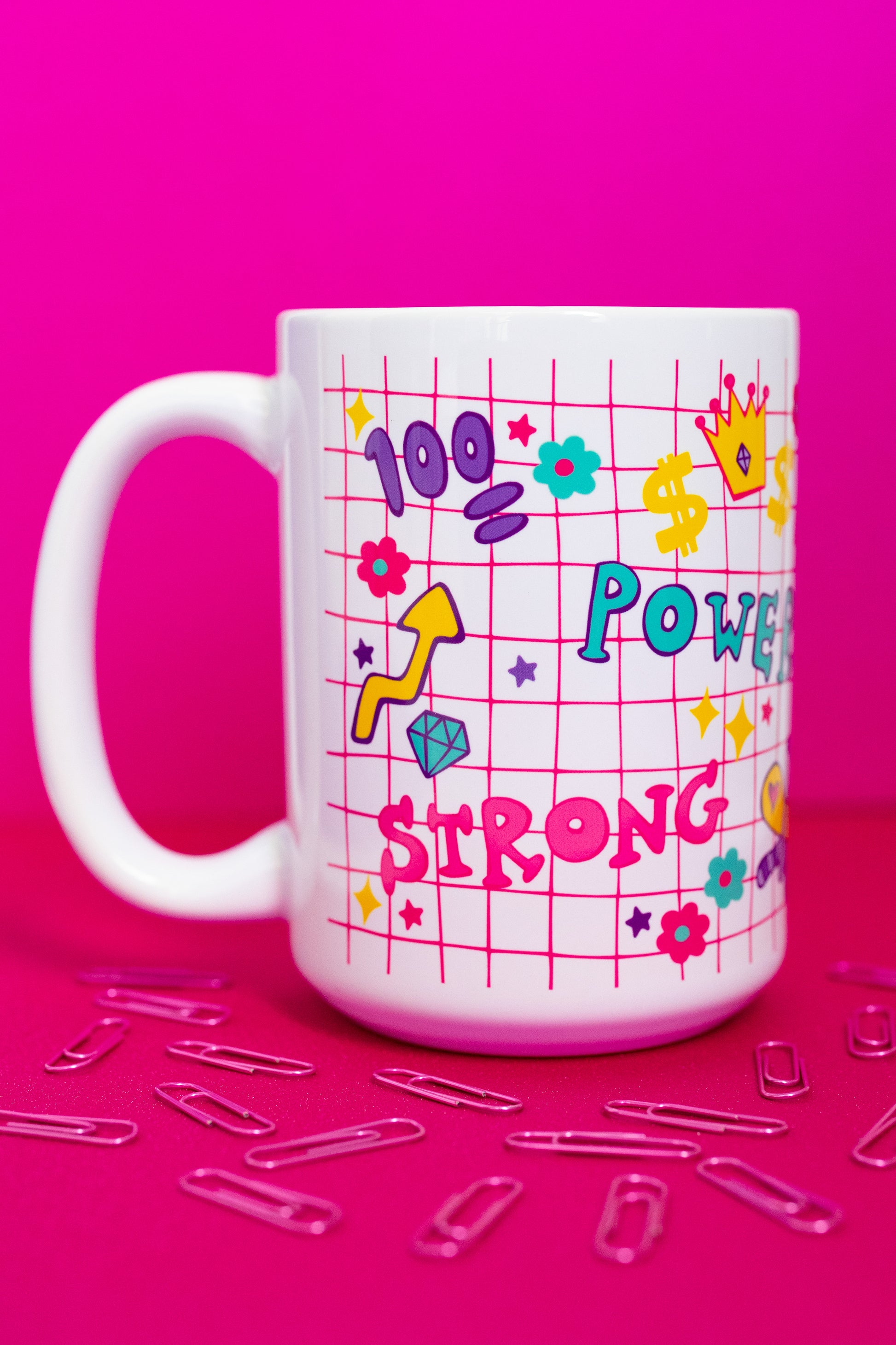 Left side of Belivra ceramic mug featuring colorful doodles and motivational words like "Power" and "Strong" on a red grid background – includes fun icons like a crown, flexing arm, dollar signs, and diamonds. Styled on a hot pink surface with scattered paperclips, this bold and empowering mug is perfect for women who love fun, energetic workspace vibes.
