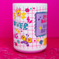 Angled view of Belivra motivational mug featuring the quote "Stay Badass. Keep Sipping." with colorful grid-style background and empowering icons like a butterfly, crown, lightning bolts, dollar signs, and hearts. Styled on a vibrant pink surface with scattered paperclips, perfect for bold women, creative workspaces, and empowering coffee moments.