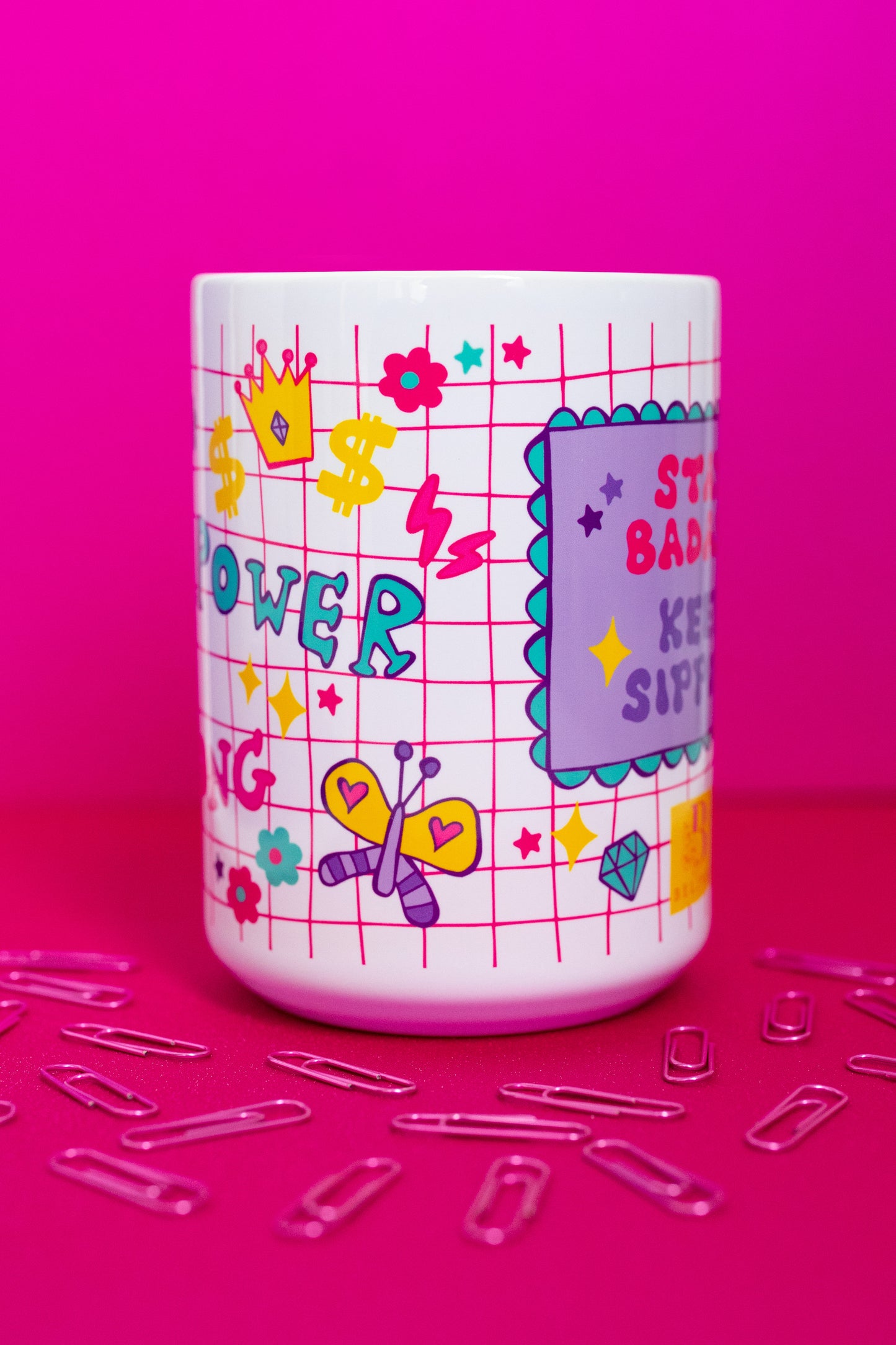 Angled view of Belivra motivational mug featuring the quote "Stay Badass. Keep Sipping." with colorful grid-style background and empowering icons like a butterfly, crown, lightning bolts, dollar signs, and hearts. Styled on a vibrant pink surface with scattered paperclips, perfect for bold women, creative workspaces, and empowering coffee moments.
