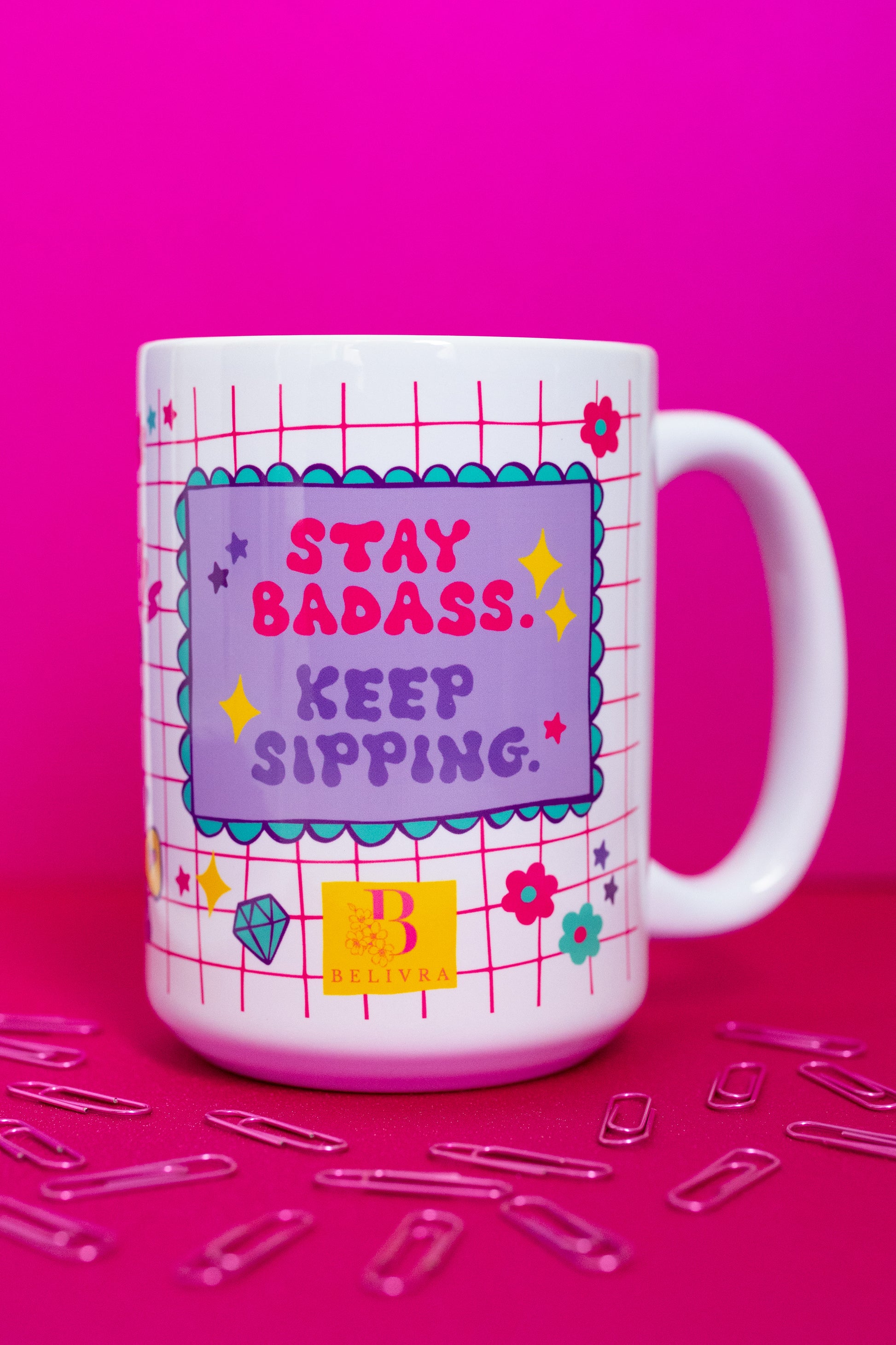 Front view of Belivra ceramic mug with empowering quote "Stay Badass. Keep Sipping." in bold red and purple text on a lavender background – set against a playful pink grid design with doodles like diamonds, flowers, and stars. Displayed on a hot pink surface with scattered paperclips, perfect for confident women, office desk vibes, and motivational coffee moments.