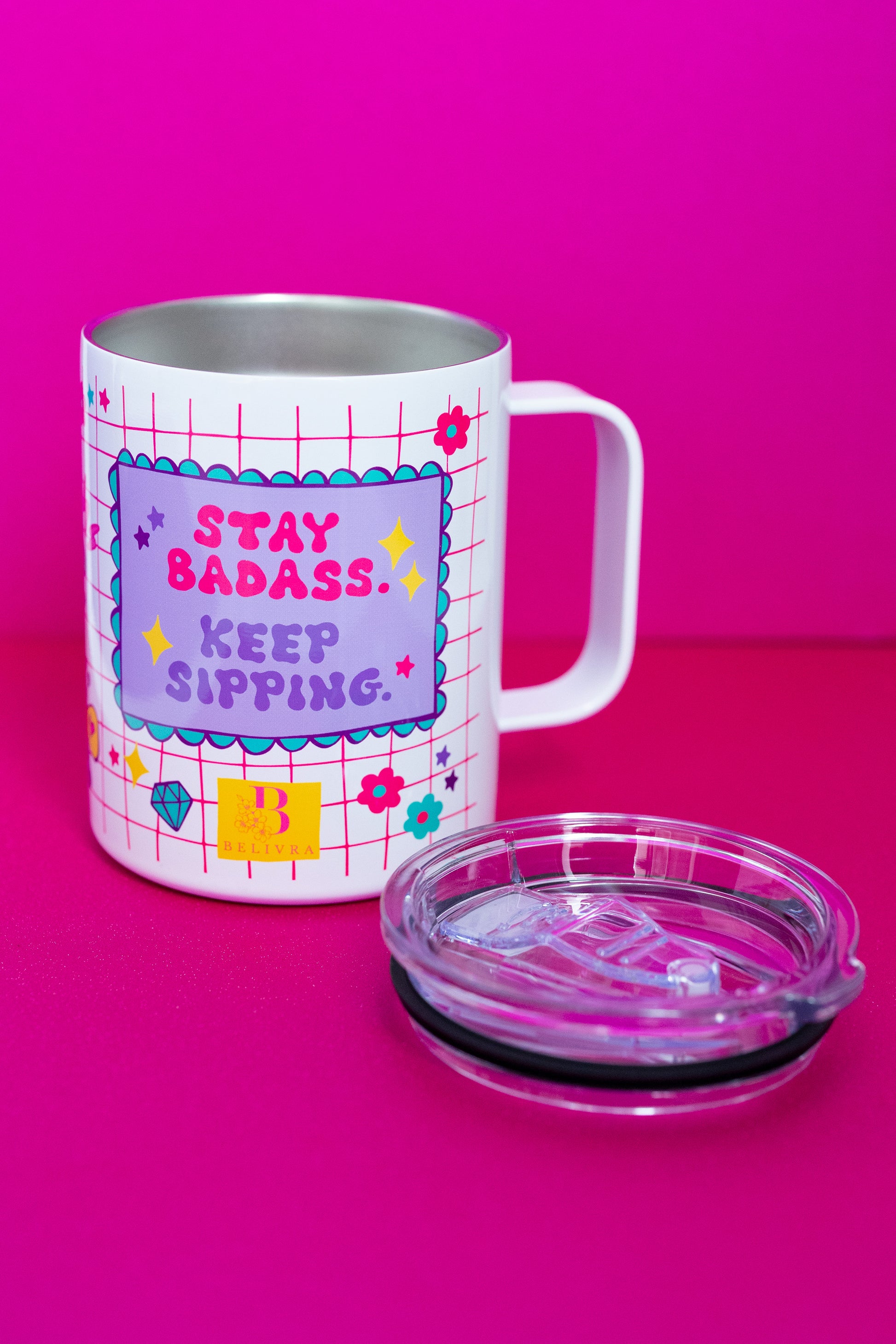 Belivra stainless steel tumbler mug with the quote "Stay Badass. Keep Sipping." in bold lettering – shown without the lid to highlight its insulated interior. Features a vibrant pink grid design with playful doodles like flowers, diamonds, and stars. Comes with a clear spill-resistant lid, perfect for hot or cold drinks, desk vibes, and empowering sips on the go.