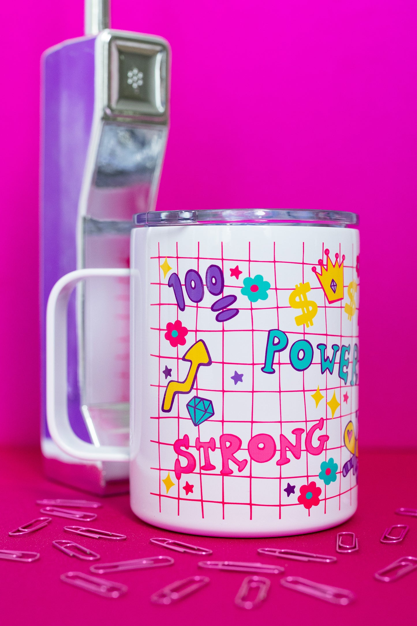 Side view of Belivra insulated tumbler mug with clear lid, featuring colorful doodles and bold words like "Power" and "Strong" on a pink grid background – decorated with symbols like a crown, flexing arm, dollar signs, and flowers. Styled on a vibrant pink surface with a retro phone and paperclips, perfect for on-the-go drinks, desk motivation, and empowering daily rituals.