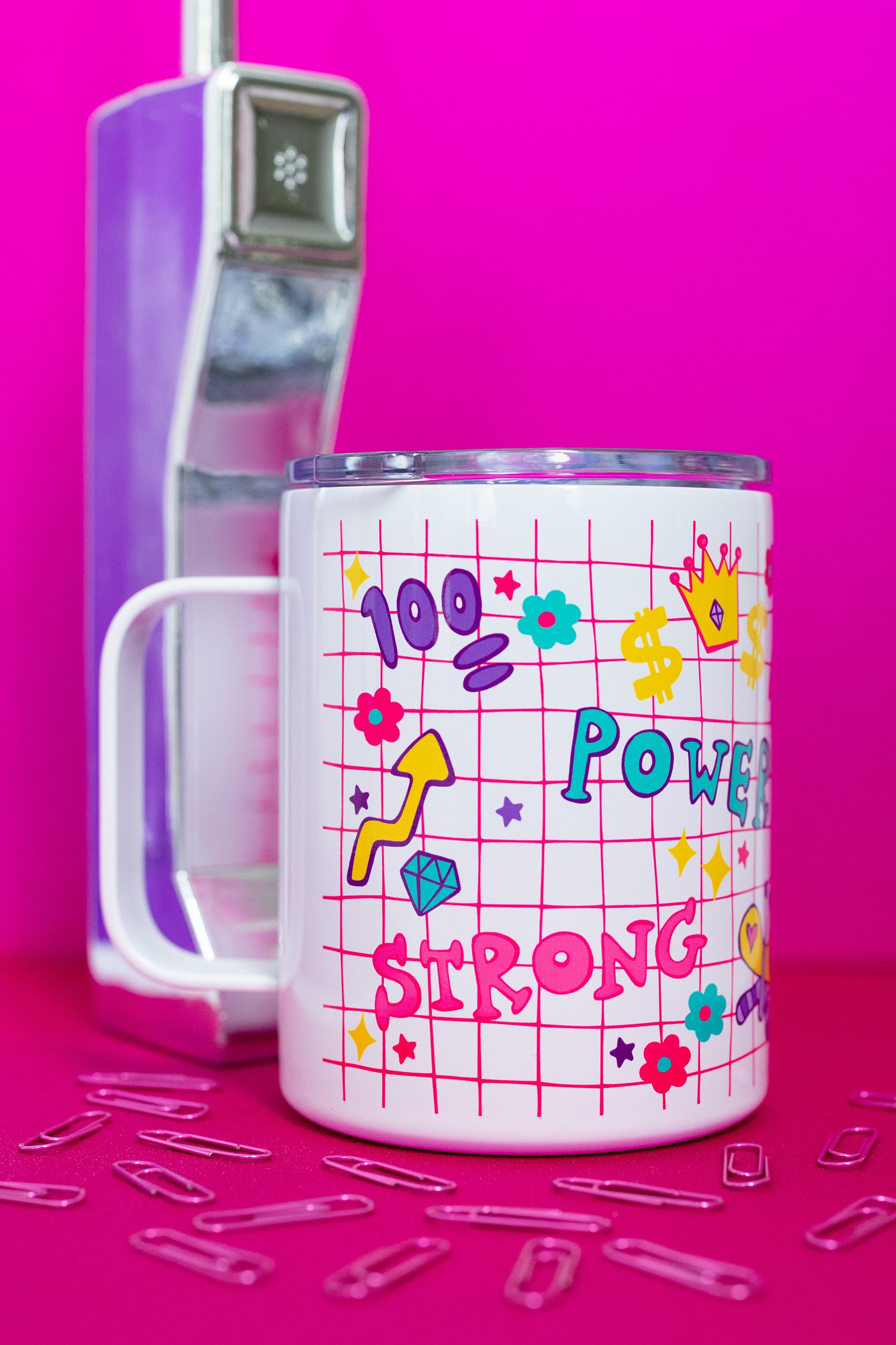 Side view of Belivra insulated tumbler mug with clear lid, featuring colorful doodles and bold words like "Power" and "Strong" on a pink grid background – decorated with symbols like a crown, flexing arm, dollar signs, and flowers. Styled on a vibrant pink surface with a retro phone and paperclips, perfect for on-the-go drinks, desk motivation, and empowering daily rituals.