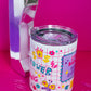 Angled view of Belivra insulated tumbler mug with clear lid, featuring the quote "Stay Badass. Keep Sipping." and colorful doodles like a butterfly, crown, lightning bolts, and dollar signs on a red grid background. Styled with a retro phone and paperclips on a hot pink surface – a bold, empowering mug perfect for busy women, coffee lovers, and motivational desk setups.