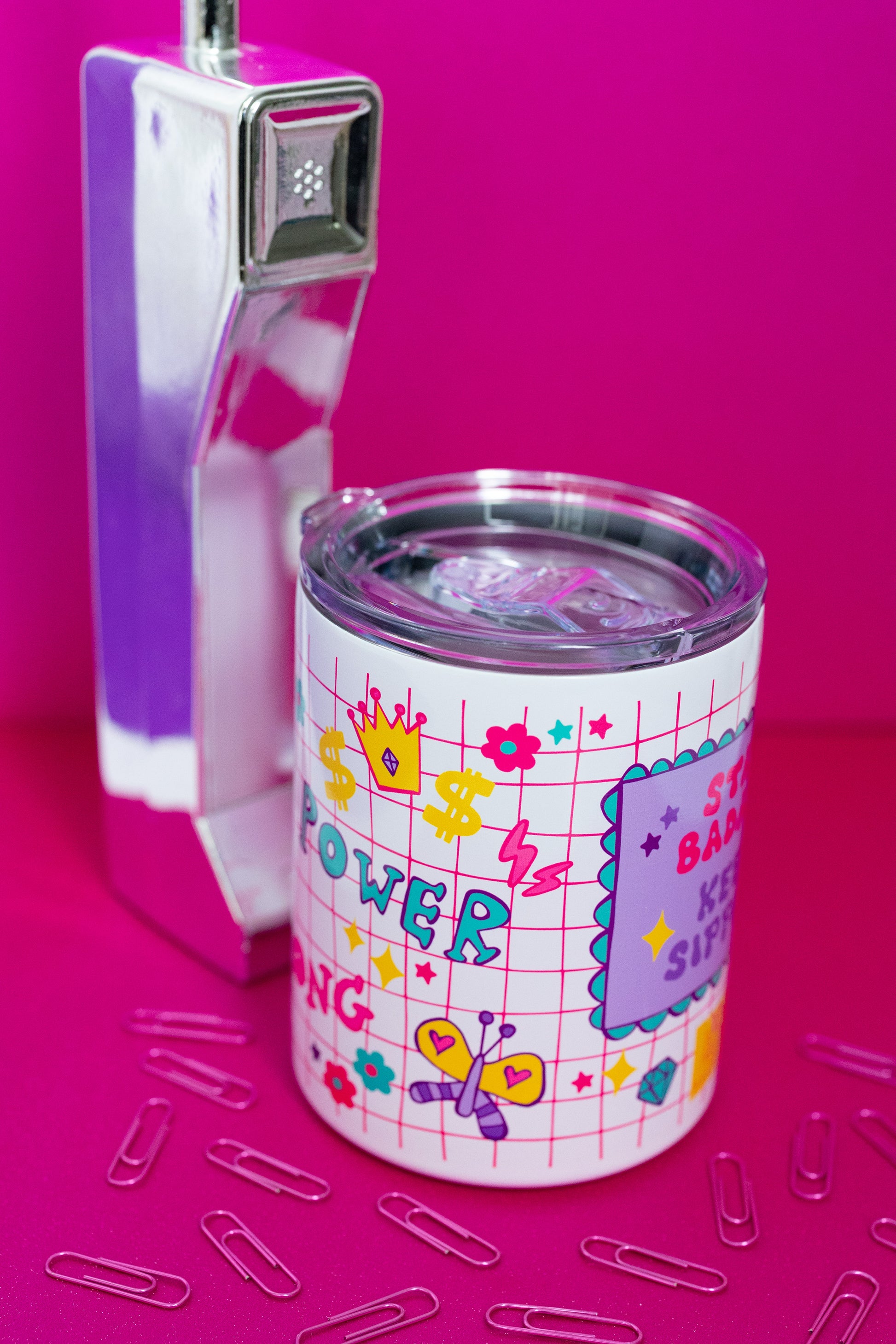 Angled view of Belivra insulated tumbler mug with clear lid, featuring the quote "Stay Badass. Keep Sipping." and colorful doodles like a butterfly, crown, lightning bolts, and dollar signs on a red grid background. Styled with a retro phone and paperclips on a hot pink surface – a bold, empowering mug perfect for busy women, coffee lovers, and motivational desk setups.