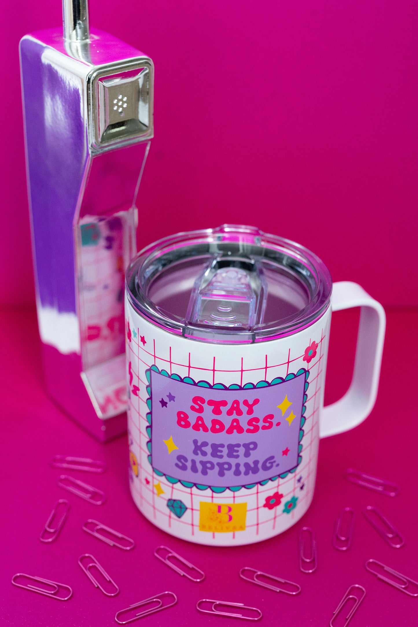 Top-down view of Belivra insulated tumbler mug with clear spill-resistant lid and bold quote "Stay Badass. Keep Sipping." in vibrant pink and purple lettering. Features a fun red grid design with colorful icons like diamonds, flowers, and stars. Styled on a hot pink surface with a retro phone and scattered paperclips, ideal for empowering coffee moments and stylish desk vibes.
