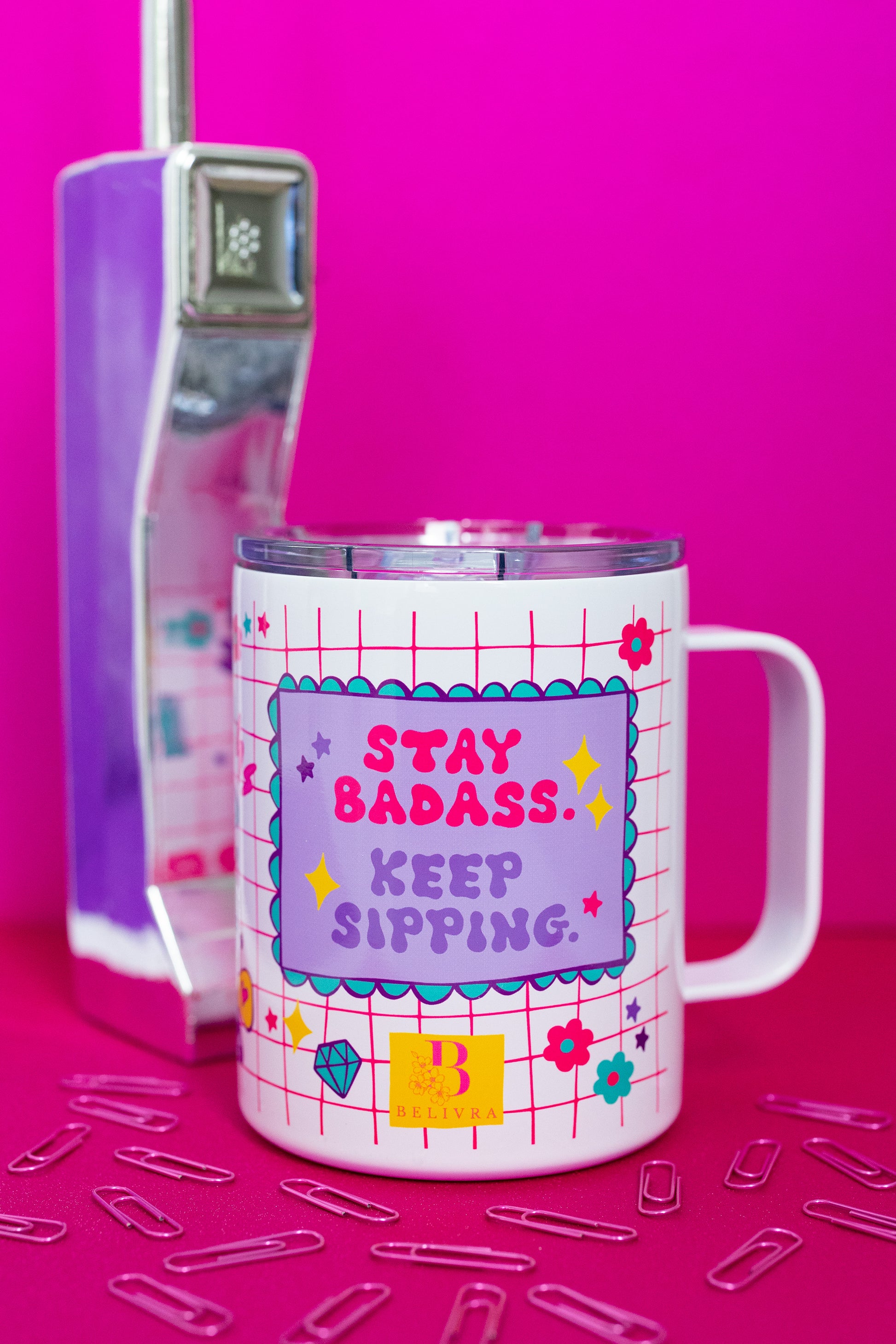 Front view of Belivra insulated tumbler mug with clear lid and bold affirmation "Stay Badass. Keep Sipping." in pink and purple text – set against a playful red grid background with doodles like flowers, diamonds, and stars. Displayed with a retro phone and scattered paperclips on a hot pink surface, this travel-friendly mug is perfect for empowering coffee or tea moments at home, work, or on the go.