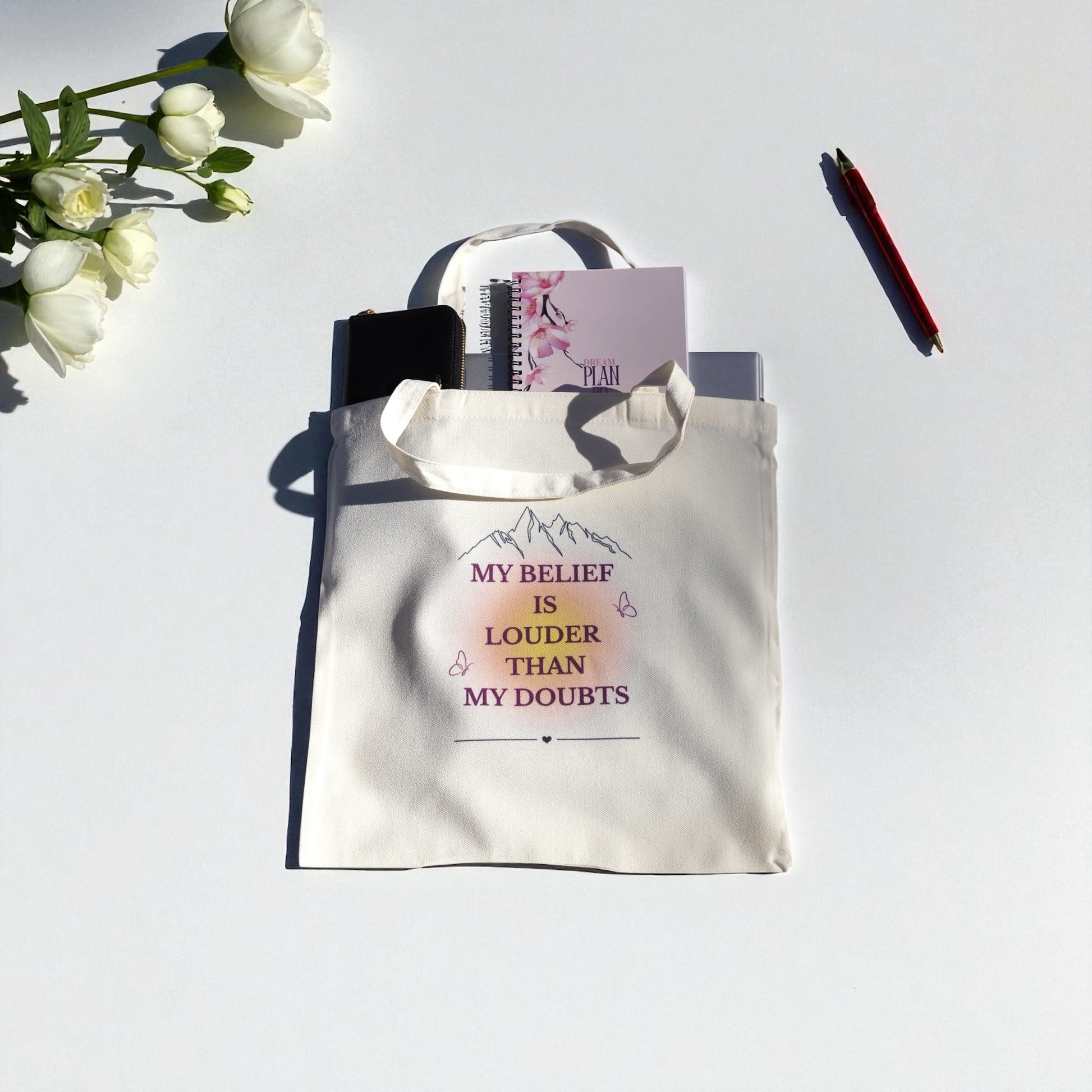 Cream tote bag with the quote “My Belief Is Louder Than My Doubts” in plum and gold text, featuring minimalist mountain and butterfly design symbolizing confidence and growth.